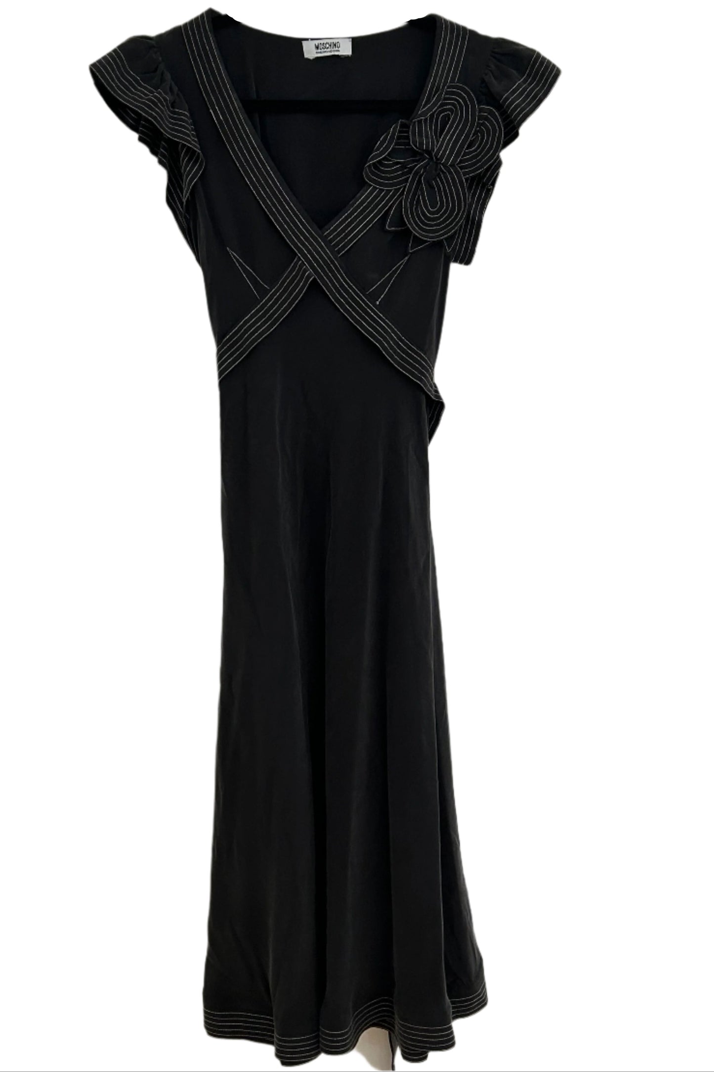 Front view of a vintage Moschino Cheap and Chic black dress with short flutter sleeves, crisscrossing ribbon details on the bodice, and a large floral applique on the left shoulder. The dress appears to have a fitted silhouette and a midi-length hem.