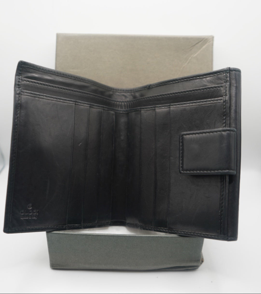 An open view of the black leather Gucci bifold wallet, showing multiple card slots on the left side and a snap closure on the right. The wallet is positioned flat with the interior visible, highlighting its minimalist design and smooth leather texture. The subtle Gucci branding is visible on the bottom left corner, emphasizing the wallet's luxury craftsmanship.
