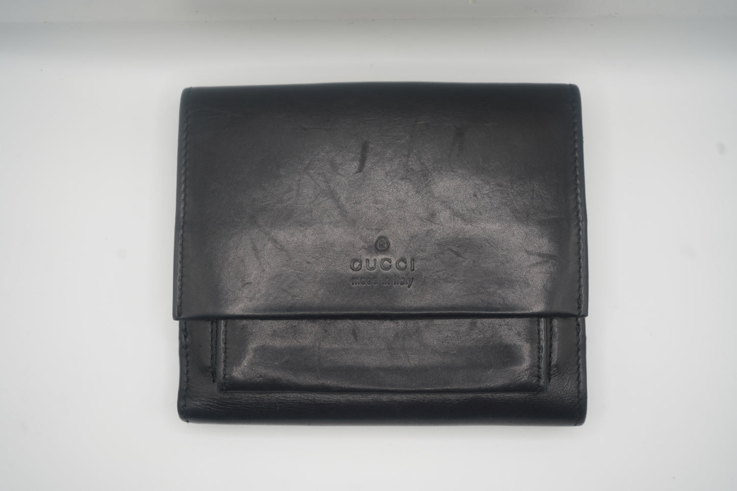 A sleek, black leather Gucci bifold wallet with subtle branding at the center. The wallet is shown from the front, highlighting its minimalist design and premium finish. The top fold is closed, and the leather has a smooth, polished appearance, suggesting both luxury and durability.