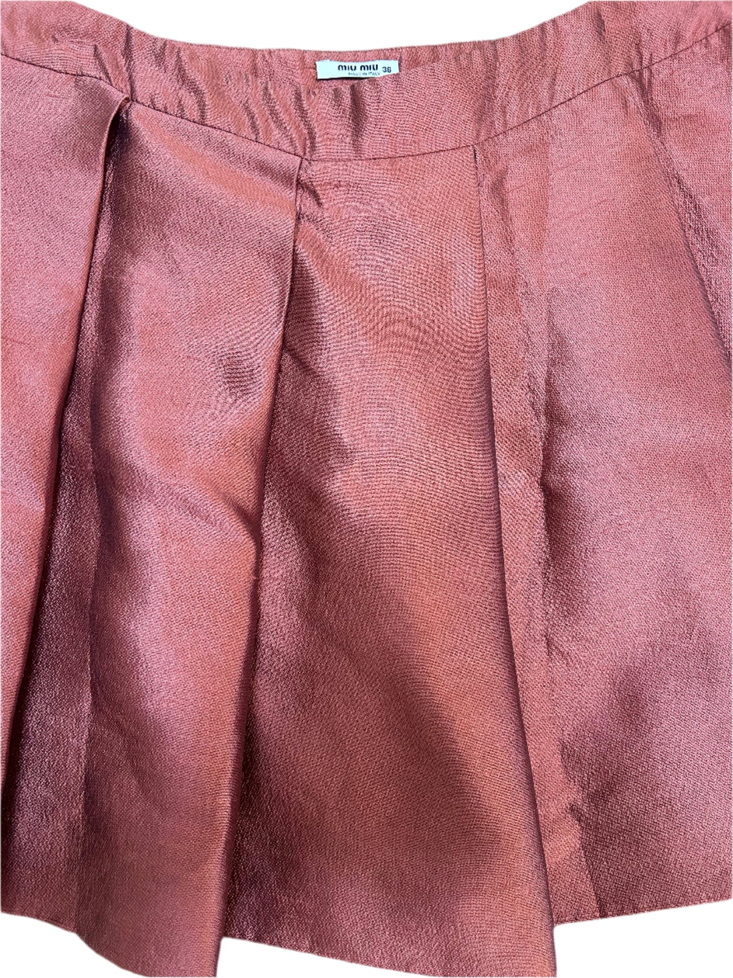 Detailed shot of a Miu Miu mini skirt in a metallic burgundy fabric with a structured silhouette. The skirt features sharp pleats for added volume and a clean waistband design