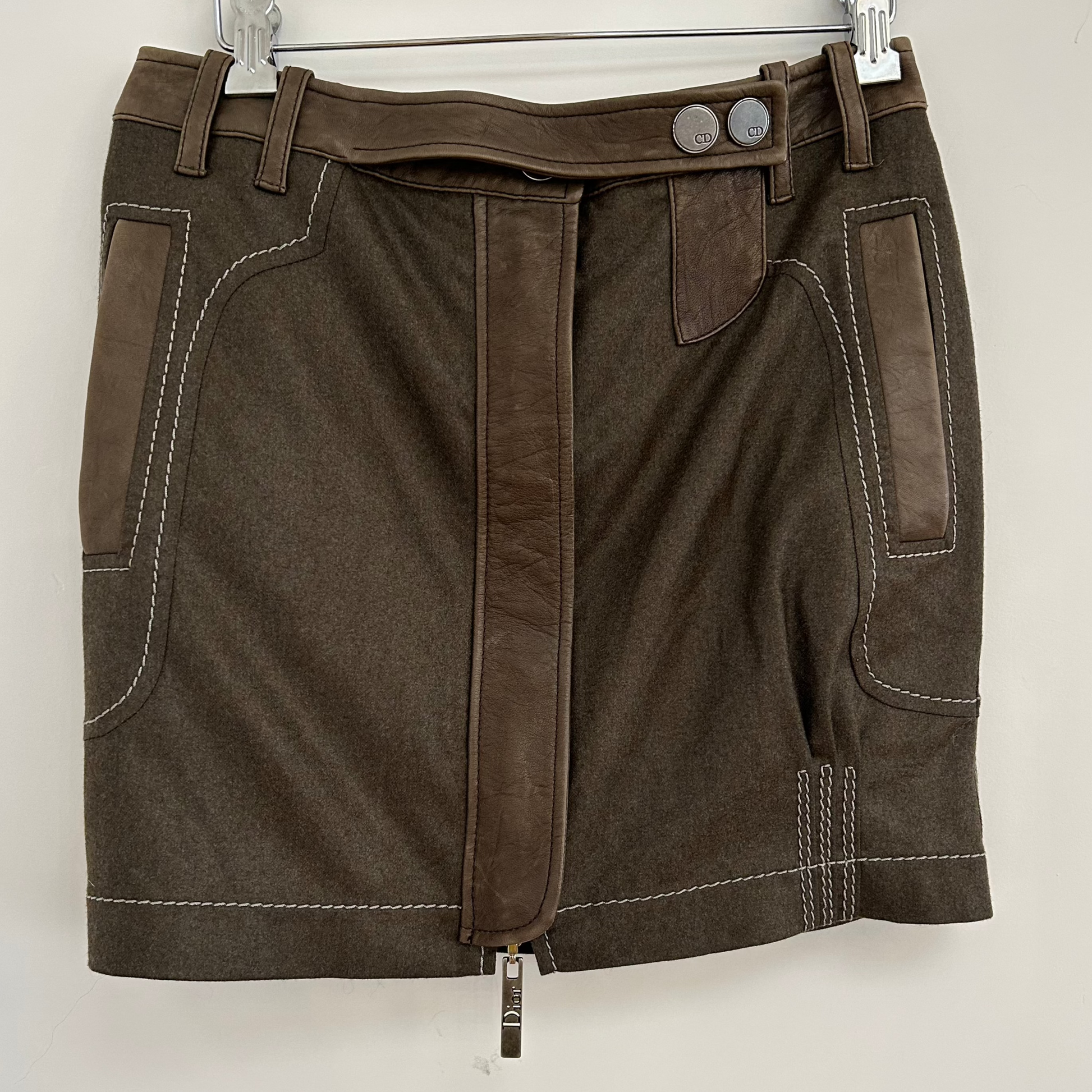 Front view of a 2000s Christian Dior wool and lambskin mini skirt in brown, featuring contrasting leather panels, white topstitching, and branded CD button details. A luxurious and edgy piece showcasing Dior's iconic craftsmanship.