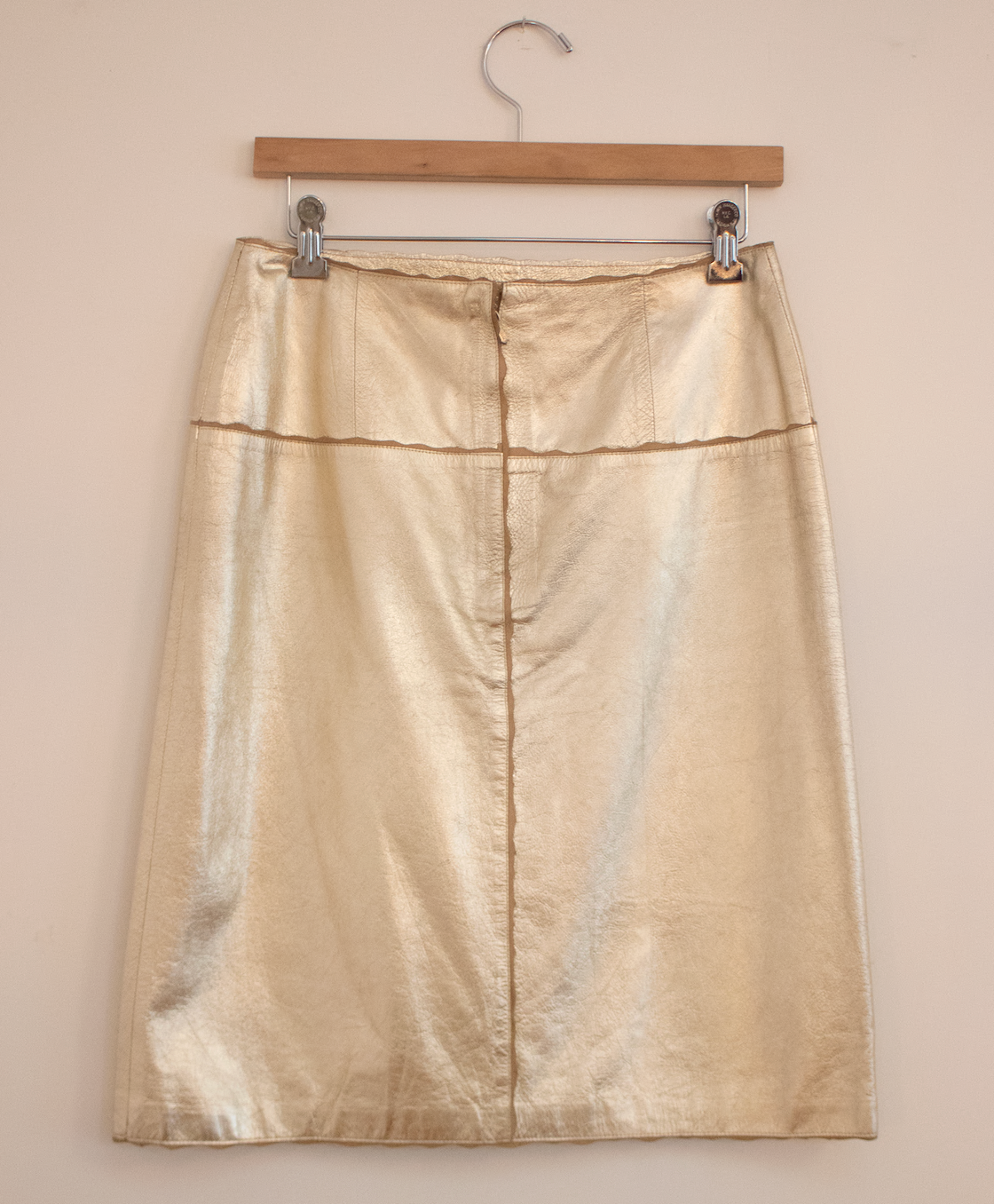 A gold-colored leather skirt with a high slit, hanging on a wooden hanger against a plain background.