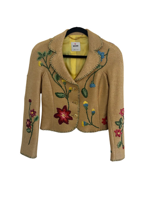 Front view of a Moschino tan woven straw blazer with a tailored design, featuring vibrant embroidered floral patterns in red, yellow, blue, and green. The blazer includes a notched collar, button-front closure, and a yellow satin lining, showcasing a playful yet sophisticated style.
