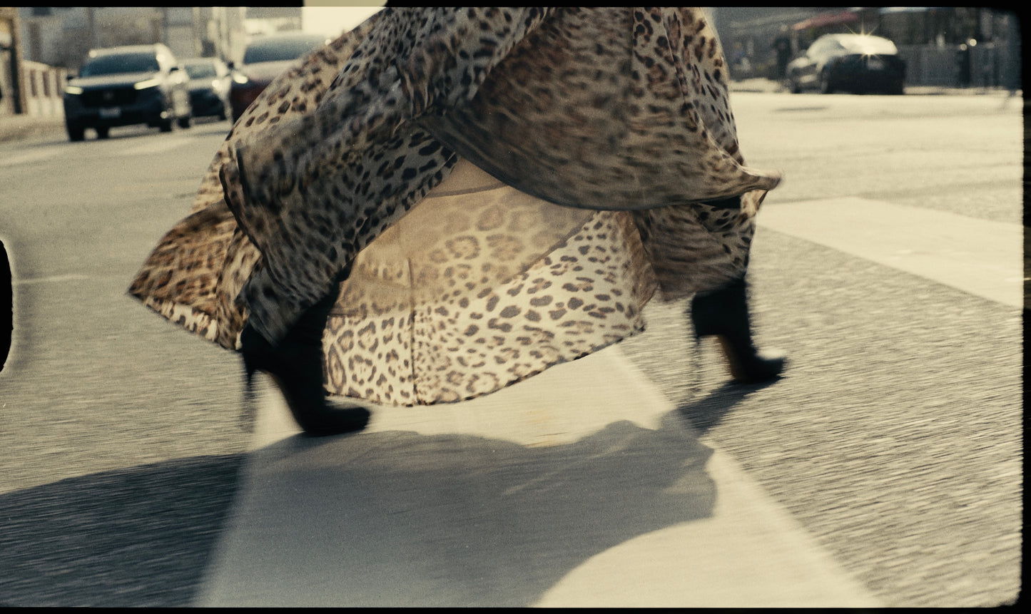 A Class Cavalli asymmetric maxi skirt in animal print, flowing gracefully as the wearer walks. The skirt's hemline is noticeably longer on one side, trailing behind, while the other side rests above the ground. The fabric ripples in motion, revealing small traces of wear at the bottom. The outfit is paired with high-heeled boots, adding a stylish touch to the flowing garment.